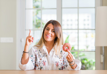 Young woman at home eating a doughnut surprised with an idea or question pointing finger with happy face, number one