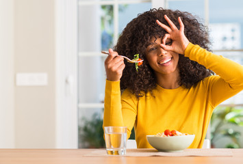 African american woman eating pasta salad at home with happy face smiling doing ok sign with hand on eye looking through fingers