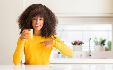 African american woman drinking orange juice in a glass with surprise face pointing finger to...