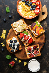Fresh breakfast - corn flakes with milk and berries, homemade waffles with fruit on a dark background. The view from above, flat lay.