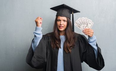 Young brunette woman over grunge grey wall wearing graduate uniform holding dollars annoyed and frustrated shouting with anger, crazy and yelling with raised hand, anger concept