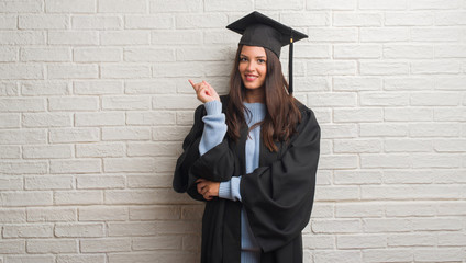 Young brunette woman standing over white brick wall wearing graduate uniform very happy pointing...