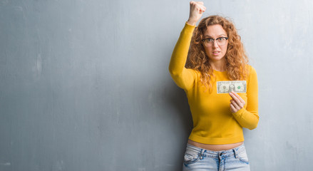 Young redhead woman over grey grunge wall holding a dollar annoyed and frustrated shouting with anger, crazy and yelling with raised hand, anger concept