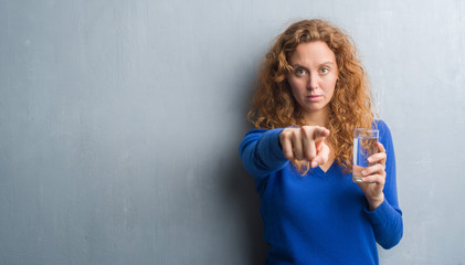 Young redhead woman over grey grunge wall drinking water pointing with finger to the camera and to...