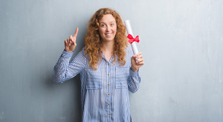 Young redhead business woman over grey grunge wall holding diploma surprised with an idea or question pointing finger with happy face, number one