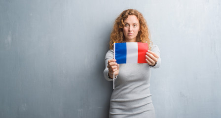 Young redhead woman over grey grunge wall holding flag of France with a confident expression on smart face thinking serious