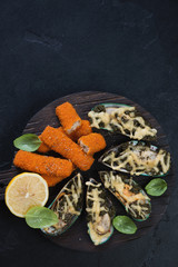 Baked mussels and fried fish sticks or breaded fish fillet, above view with space on a black stone background, vertical shot