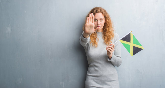 Young redhead woman over grey grunge wall holding flag of Jamaica with open hand doing stop sign with serious and confident expression, defense gesture