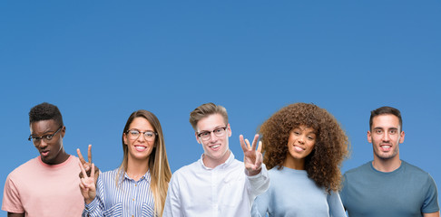 Composition of group of friends over blue blackground showing and pointing up with fingers number two while smiling confident and happy.