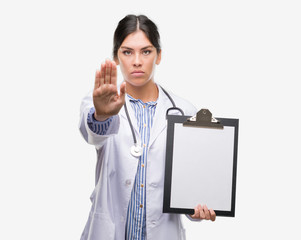 Young hispanic doctor woman holding clipboard with open hand doing stop sign with serious and...