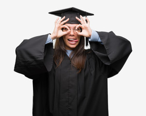 Young hispanic woman wearing graduated cap and uniform doing ok gesture like binoculars sticking tongue out, eyes looking through fingers. Crazy expression.