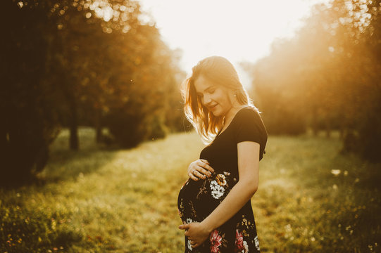 Young beautiful pregnant woman standing in sunlight in park at sunset
