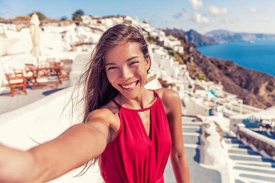 Travel vacation tourist girl selfie photo with phone on Santorini holiday. Happy asian woman in Oia smiling taking self-portrait picture with smartphone on summer vacation in Greece destination.