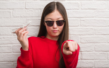 Young Chinese woman over brick wall holding plane toy pointing with finger to the camera and to...