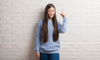 Young Chinise woman over white brick wall pointing finger up with successful idea. Exited and happy. Number one.