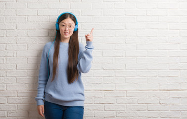 Young Chinese woman over brick wall wearing headphones surprised with an idea or question pointing...
