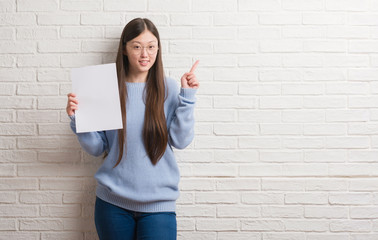 Young Chinese woman over brick wall holding blank paper surprised with an idea or question pointing...