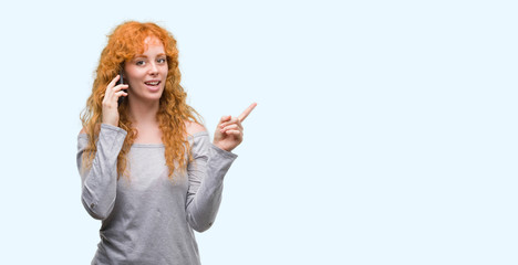 Young redhead woman talking on the phone very happy pointing with hand and finger to the side