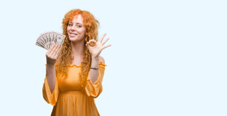 Young redhead woman holding dollars doing ok sign with fingers, excellent symbol