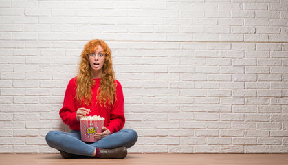 Young redhead woman sitting over brick wall eating popcorn scared in shock with a surprise face,...