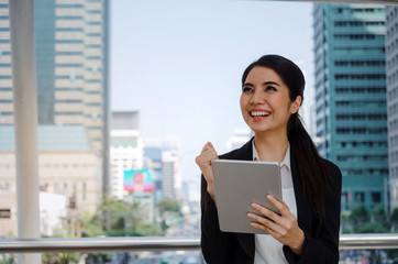 beautiful happy young asian business woman smiling wearing modern black suit cheer up hand with mobile tablet in building city background, network technology, internet, financial, investment concept