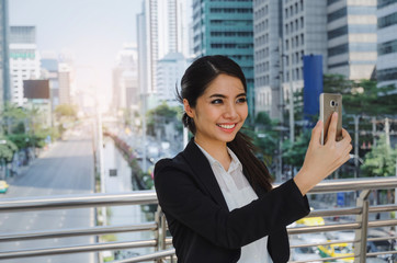 young asian business woman smiling wearing modern black suit video conference with mobile phone in building city background, network technology, internet, digital financial and investment concept