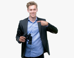 Young handsome blond man looking through binoculars with surprise face pointing finger to himself
