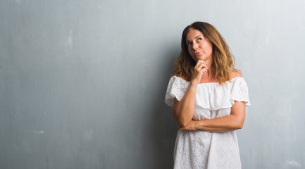 Middle age hispanic woman standing over grey grunge wall with hand on chin thinking about question, pensive expression. Smiling with thoughtful face. Doubt concept.