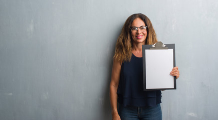 Middle age hispanic woman standing over grey grunge wall holding clipboard with a happy face standing and smiling with a confident smile showing teeth