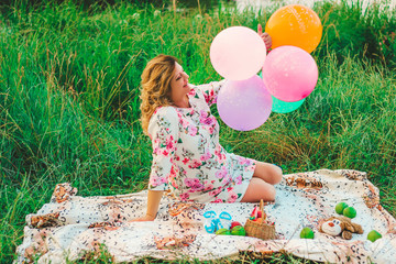 Young beautiful pregnant woman sitting at a picnic on a blanket in the park with colorful balloons in hands closeup