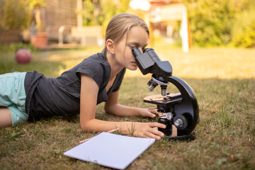 Ein 9 jähriges Mädchen liegt auf dem Rasen im Garten und schaut in das Okular eines Mikroskops. Vor ihr liegt ein Blatt Papier