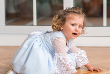 Fototapeta premium Beautiful blonde child in a dress crying with a trantrum sitting on the floor at home