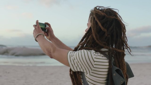 young carefree man with dreadlocks taking photos on beach using phone wearing stripe shirt