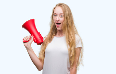 Blonde teenager woman holding megaphone scared in shock with a surprise face, afraid and excited with fear expression