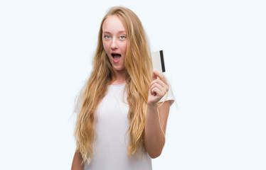 Blonde teenager woman holding credit card scared in shock with a surprise face, afraid and excited with fear expression