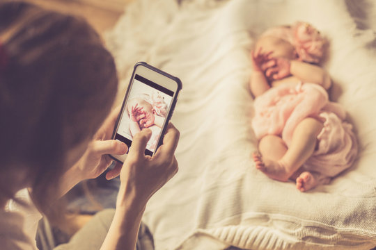 A woman holding a smartphone and takes a picture of a newborn baby girl sleeping on a white blanket. Selective focus.