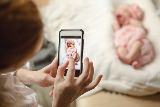 A woman holding a smartphone and takes a picture of a newborn baby girl sleeping on a white blanket. Selective focus.