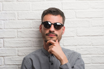 Young adult man wearing sunglasses standing over white brick wall serious face thinking about...