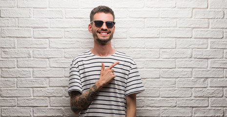Young adult man wearing sunglasses over white brick wall cheerful with a smile of face pointing with hand and finger up to the side with happy and natural expression on face looking at the camera.