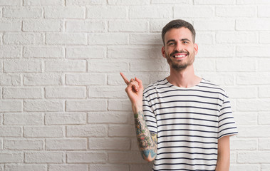 Young adult man standing over white brick wall very happy pointing with hand and finger to the side