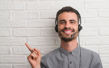 Young adult man over brick wall working as operator very happy pointing with hand and finger to the...