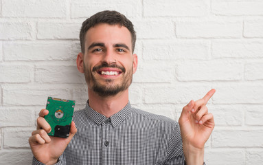 Young adult man over brick wall holding hard drive very happy pointing with hand and finger to the...