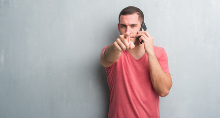 Young caucasian man over grey grunge wall talking on the phone pointing with finger to the camera and to you, hand sign, positive and confident gesture from the front