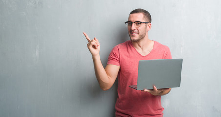 Young caucasian man over grey grunge wall using computer laptop very happy pointing with hand and...