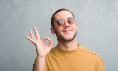 Fototapeta premium Young caucasian man over grey grunge wall wearing sunglasses doing ok sign with fingers, excellent symbol