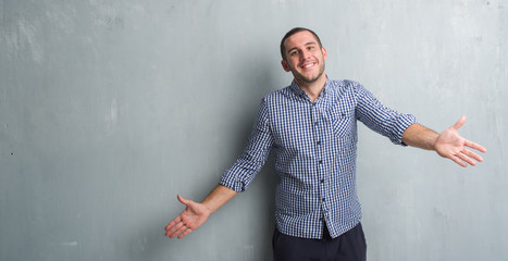 Young caucasian man over grey grunge wall looking at the camera smiling with open arms for hug. Cheerful expression embracing happiness. - obrazy, fototapety, plakaty