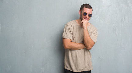 Young caucasian man over grey grunge wall wearing sunglasses looking confident at the camera with smile with crossed arms and hand raised on chin. Thinking positive.