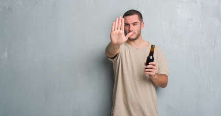 Young caucasian man over grey grunge wall holding bottle beer with open hand doing stop sign with serious and confident expression, defense gesture