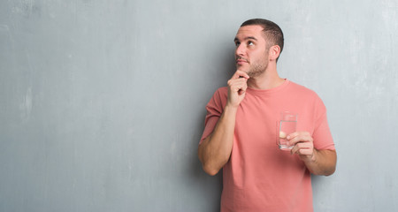 Young caucasian man over grey grunge wall drinking a glass of water serious face thinking about...