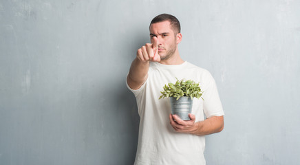 Young caucasian man over grey grunge wall holding plant pot pointing with finger to the camera and...
